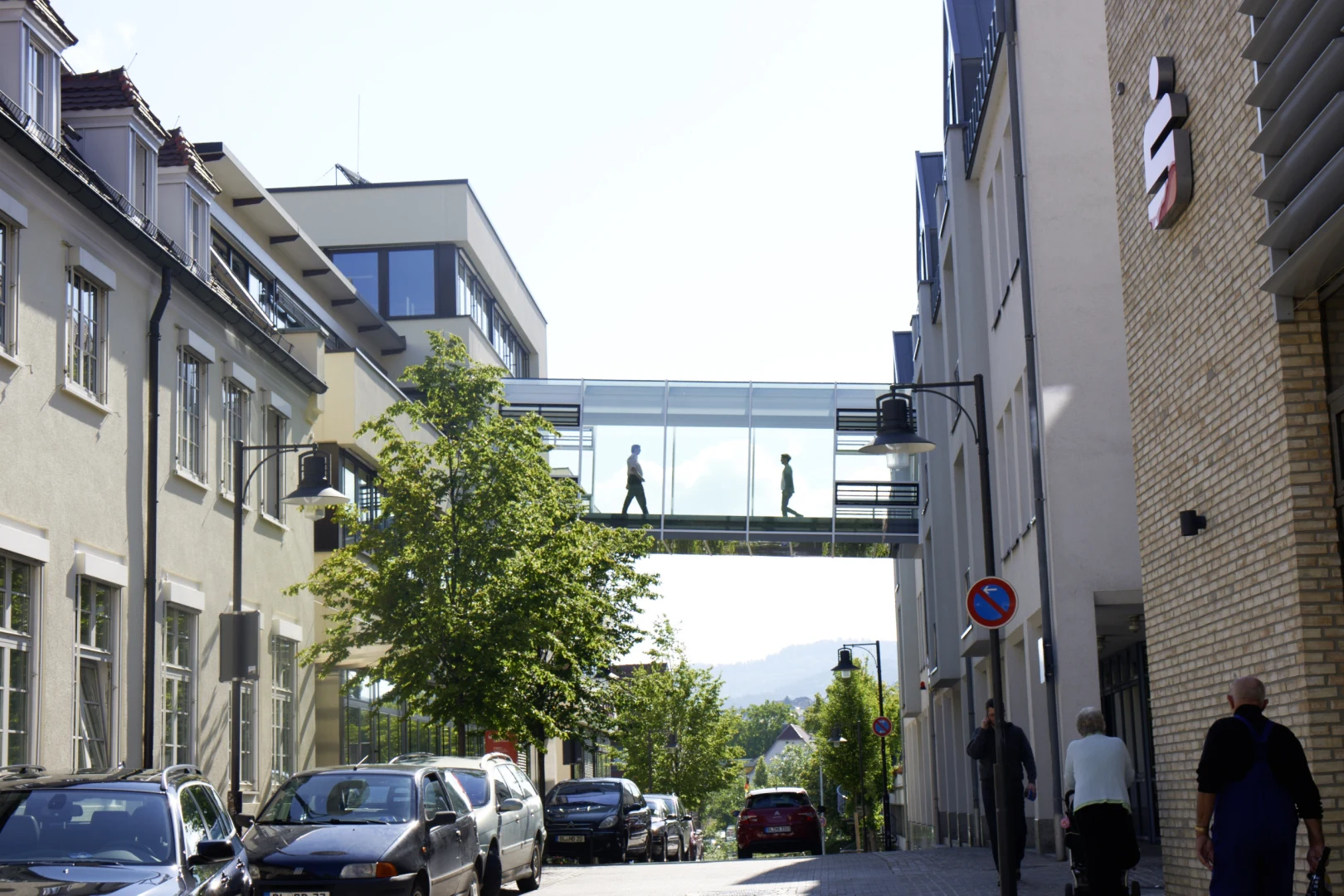 Man sieht die Hechinger Str. in Balingen, mit Bäumen und parkenden Autos. Über die Straße geht ein gläserner Übergang. In diesem Übergang treffen sich zwei Personen.